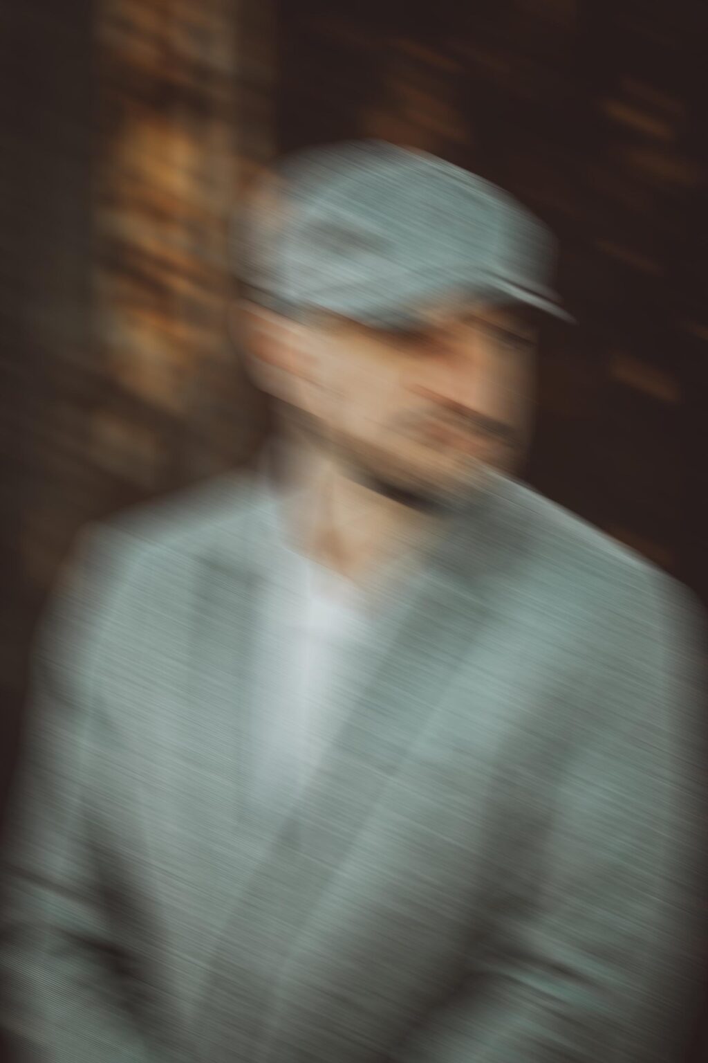 Image floue d'un homme portant une casquette et un costume gris, rappelant le style avant-gardiste souvent observé chez le photographe nu de Lyon. Son visage et sa silhouette sont obscurcis par le flou de mouvement, ce qui rend les traits et les détails du visage indiscernables. L'arrière-plan est composé de couleurs et de formes sombres et indistinctes.