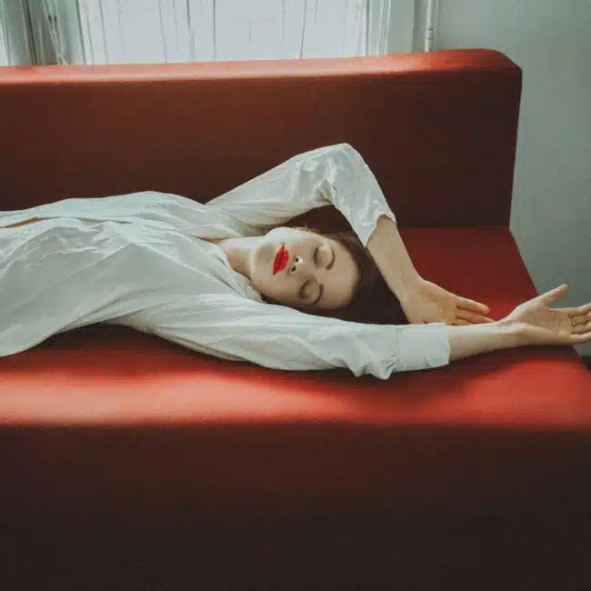 Une personne avec du rouge à lèvres rouge et une chemise blanche est allongée sur un canapé rouge, les bras tendus au-dessus de la tête, les yeux fermés, dans une pose détendue près d'une fenêtre avec une lumière naturelle douce pendant une prise de vue par un photographe professionnel.