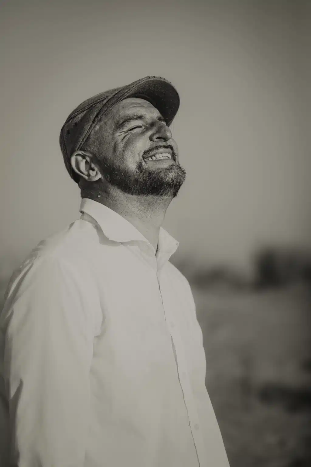 Un homme portant une chemise claire et une casquette plate se tient à l'extérieur, à Lyon, avec un large sourire et les yeux fermés, dans une photo de prise de vue en noir et blanc. L'arrière-plan flou ajoute du charme à l'instant.